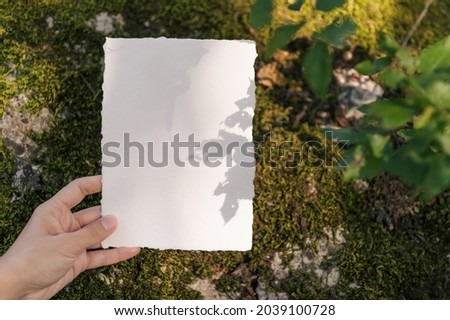 Hand holding blank paper sheet cards with empty copy space on old stone with sunlight shadows on dark background. Wedding invitation mockup template, greeting card, business card.