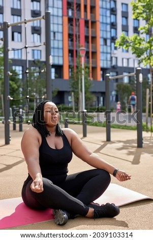 Yoga, mindfulness, healthy lifestyle concept. Young fat african woman meditating outdoors on sports ground, keep calm, breathing correctly, with eyes closed and crossed legs, on fitness mat.