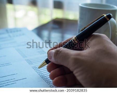Close up of a man's right hand holding a black pen to sign off or checking document with background of a white mug on a wooden table. Selective focus. Working from home concept. Kontrak dokumen, pajak