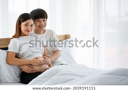 Portrait of a good-looking young Asian man and woman wearing a white nightdress sitting on a bed together. They are smiling happily and touching the belly of a pregnant mother.
