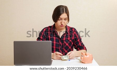 Serious young woman in checkered shirt sits at table near laptop planning family budget closeup