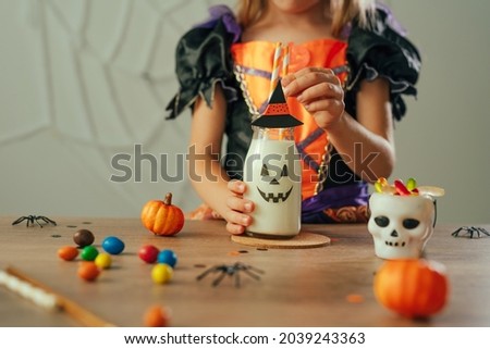 Close up of little girl dressed Halloween witch costume sitting at the table with milkshake in a glass bottle decorated with jack-o-lantern face indoor. Selective focus.
