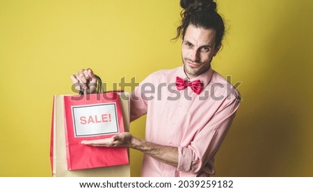 Isolated Young Caucasian Personal Shopper on a Yellow Colored Background Holding Multiple Gift Bags Pointing to the  Sign Sale On It. Wearing a Pink Shirt With a Red Bow Tie and a Man Bun.
