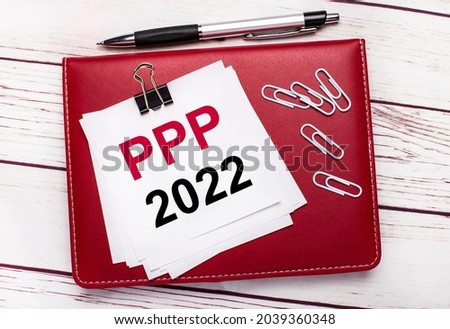 On a light wooden background, a burgundy pen and notebook. On the notebook has white paper clips and white paper with the text PPP 2022. Business concept