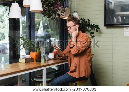 young man in glasses adjusting wireless earphones and using smartphone in cafe