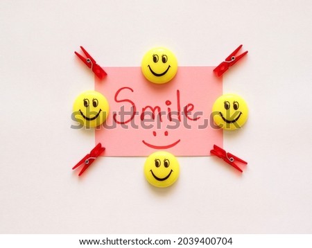 Smile word on white background, on pink card, on table. Red mini craft clothes pegs and smile face fridge magnets. Top view, flat lay.
