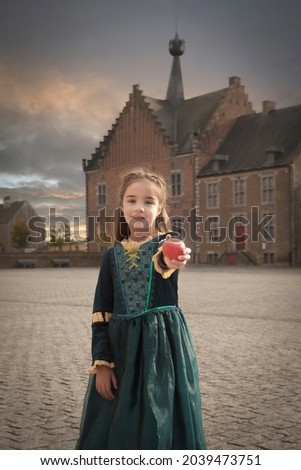 A little girl in a medieval dress against the background of a castle stretches out a red apple