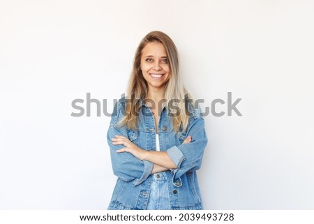A young caucasian smiling charming pretty blonde woman in a denim jacket and jeans stands crossing her arms on her stomach isolated on a white background. Closed pose