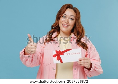 Young redhead chubby overweight woman 30s with curly hair wear pink shirt casual clothes hold gift certificate coupon voucher card for store show thumb up gesture isolated on pastel blue background