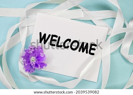 The greeting is written on a white sheet which lies on a blue background next to a white ribbon and a purple flower.