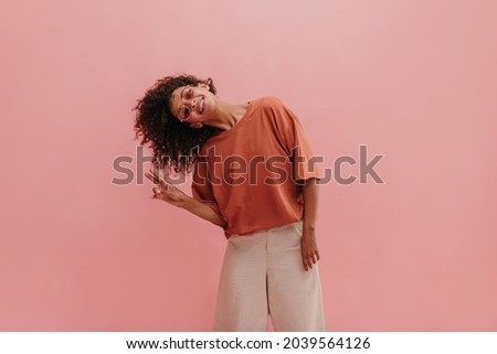 impressive, funny lady in background of studio background tilted head to one side, showing two thumbs up. dark short curly hair flying beautifully in photo. girl in glasses smiles broadly with teeth.