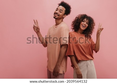 two african american friends show peace sign on pink studio background. guy and girl with dark afro hair are smiling at camera dressed in casual light-colored clothes.