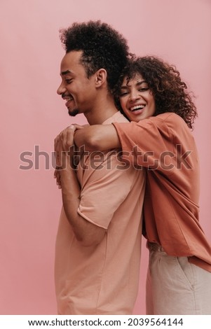 profile photo of friendly people of different genders on isolated light pink background. cute brunette with lush hair smiles broadly with teeth and hugs guy tightly from behind.