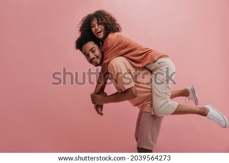 side profile full length cheerful positive cute couple in casual wear. african american man carrying woman smiling with teeth over isolated pastel pink background.