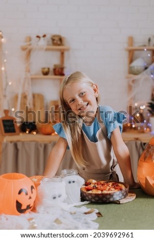 a blonde baby girl with long hair in an apron in the kitchen, decorated with pumpkins and garlands for Halloween, prepares a focaccia pie. space for text. High quality photo