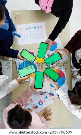 Businesspeople sitting together in office over working desk with colorful graph and charts pater, hold smartphones in different model with blank screens. Taken from top view angle.