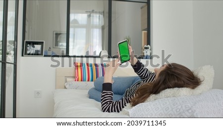 Young Woman at Home Lying on a bed using with Green Mock-up Screen Smartphone. Girl Using Mobile Phone, Browsing Internet, Watching Content, Video , Blog