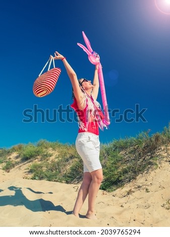 Middle aged happy woman in red swimsuit and white shorts having fun enjoying beautiful sandy beach on the sunny weather on island. Woman with an inflatable hare and a bag goes to the sea