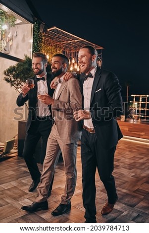 Full length of three handsome men in suits and bowties drinking whiskey and communicating Royalty-Free Stock Photo #2039784713