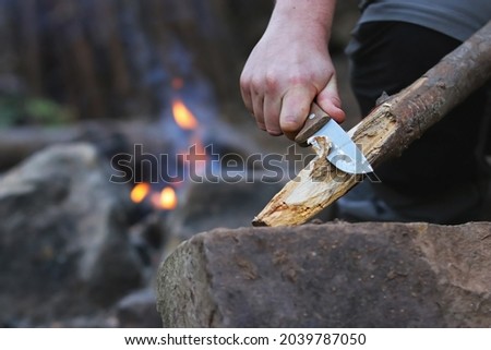 Close up of the hands of a man carving off timber to lit a fire, camp fire on the background. Hand holding knife cutting a wooden stick. Bushcraft and outdoors survival activities concept.  Royalty-Free Stock Photo #2039787050