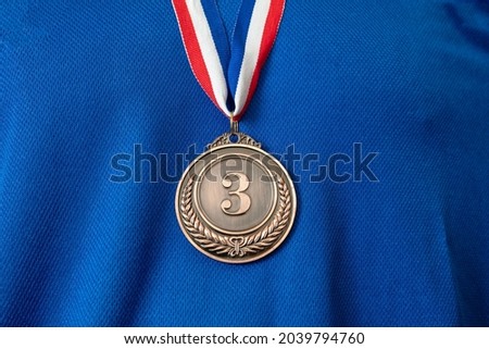 Medal bronze. Winner prize award hanging with red blue color ribbon on athlete chest. Trophy in sport for third place champion on blue color shirt background Royalty-Free Stock Photo #2039794760