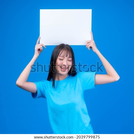 beautiful Asian young woman with bangs hair style in blue t shirt smiling and holding a blank board empty space for advertising banner , white board a blank banner isolated on blue background