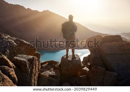 Silhouette of Man Hiker tourist man with backpack standing on the rock against beautiful sunset at lake in the mountain valley. Outdoor and trekking concept.