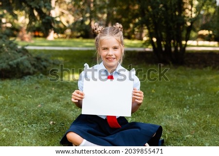 Cute girl holding a white sheet. Blonde young girl with a white sheet of paper. Little schoolgirl is holding a blank sheet.