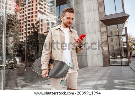 Focused millennial blonde guy during smartphone posting liking streaming standing on urban street background students communicating via cellphone gadget, hipster skateboarder holding longboard
