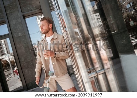Portrait of blonde young man holding cup of coffee cappuccino takeaway standing on urban street glass building background hipster skateboarder holding longboard. Extreme leisure concept. Big city life