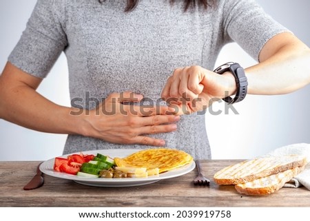 Intermittent fasting concept with a woman sitting hungry in front of food and looking at her watch to make sure she breaks fast on the correct time. A dietary modification for healthy lifestyle. Royalty-Free Stock Photo #2039919758