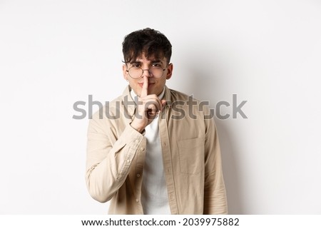 Cheerful handsome man in glasses share a secret, making surprise, lean at camera with hush gesture and gentle smile, telling to be quiet, show taboo sign, white background