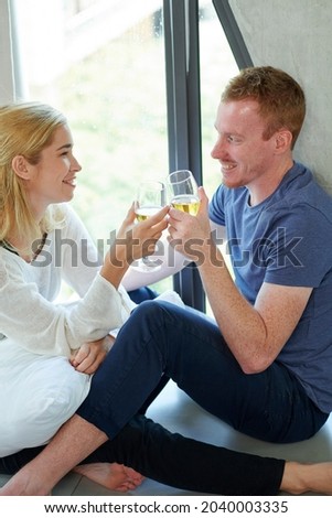 Young boyfriend and girlfriend sitting on window sill with champagne glasses in hands and enjoying romantic date