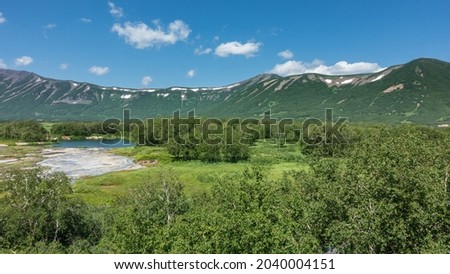 The caldera of an extinct volcano is surrounded by a mountain range.  In the valley there is a blue lake, a green meadow, thickets of deciduous trees. Clouds in the azure sky. Summer. Kamchatka. Uzon