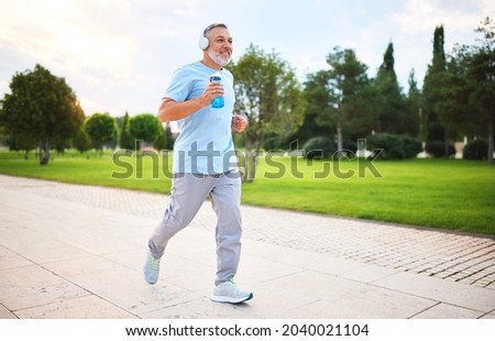Full-length photo of happy senior man in headphones and sportswear jogging running outside in city park with water bottle in hand. Healthy active lifestyle on retirement and sport outdoor concept