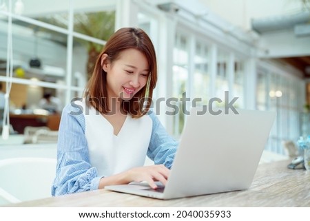 A beautiful woman wearing an Asian white shirt is sitting in front of a laptop computer shopping online with a happy smile in a bakery shop.