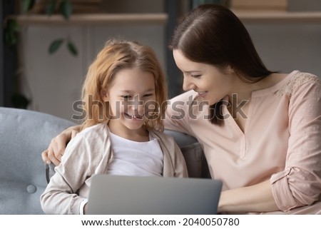 Caring young mother teaching little cute kid daughter using computer software apps, resting on comfortable sofa at home. Addicted to modern technology bonding two generations family playing online.