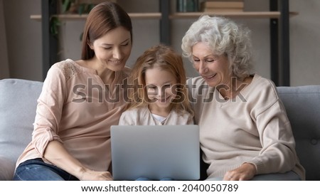 Happy bonding small kid girl using computer with caring mother and joyful middle aged retired granny, watching video photo online, relaxing together on comfortable couch, tech addiction concept.
