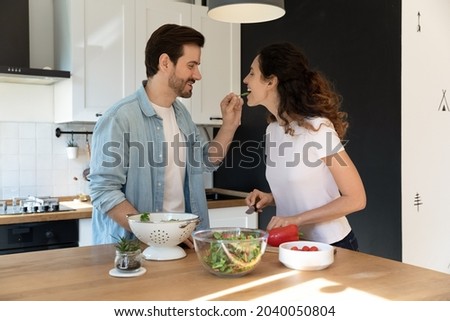 Caring husband feed beloved wife, Latina wife tasting salad leaf, married couple enjoy cook in modern kitchen at home on weekend. Romantic date, family cooking healthy vegetarian food together concept Royalty-Free Stock Photo #2040050804