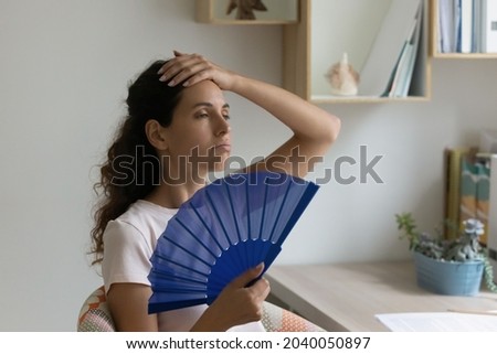 Young sweaty thirsty woman sit indoor at desk feels overheated due unbearable summer hot day, holding handheld blue fan cooling herself. Apartment without air-conditioner, need climate control concept Royalty-Free Stock Photo #2040050897