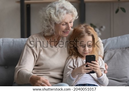 Affectionate caring middle aged grandmother cuddling small cute grandchild girl, enjoying using cellphone applications together, playing mobile games or spending time online, tech addiction concept.