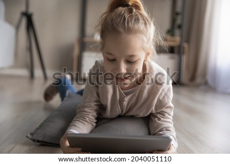 Happy adorable little schoolgirl child using digital computer touchpad tablet, lying on cozy soft pillow on warm wooden floor, enjoying playing online videogame, kids and modern tech addiction concept