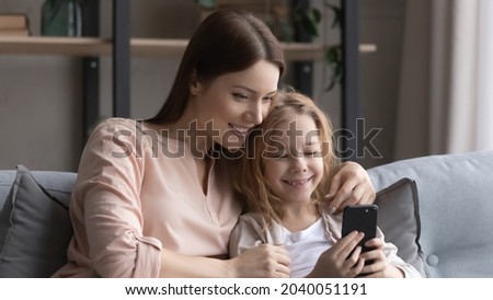 Happy bonding young mother and little child daughter using cellphone, resting together at home, enjoying playing mobile games, choosing goods shopping in internet store or using funny applications.