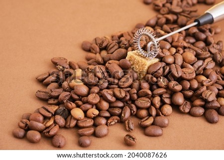 a pile of coffee beans, sugar cube and metal handheld milk steamer on brown background. Handheld frother or foam maker close up. copy space