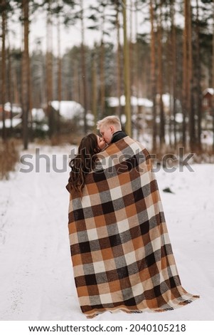A happy married couple in love is hiding under a plaid blanket, warming up while walking traveling in the winter forest in nature in cold weather, selective focus