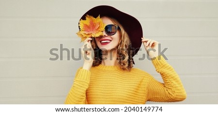 Autumn portrait of beautiful happy smiling young woman covering her eyes with yellow maple leaves wearing a knitted sweater, hat on gray background