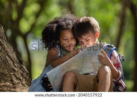 African American ethnicity boy and girl children wearing plaid shirt and backpack sitting at tree base talking and looking down on the map travel on hand in the park day light background 