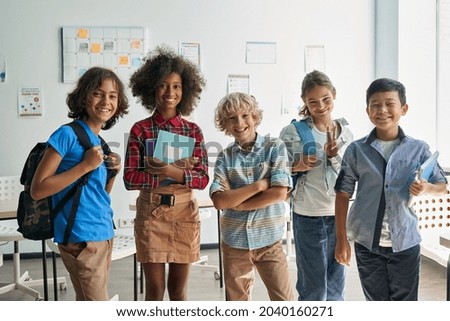 Portrait of cheerful smiling diverse schoolchildren standing posing in classroom holding notebooks and backpacks looking at camera happy after school reopen. Diversity. Back to school concept. Royalty-Free Stock Photo #2040160271