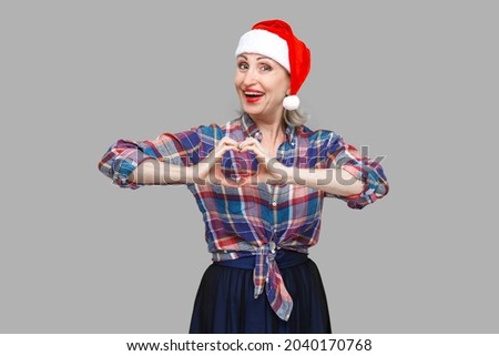 Portrait of peacefulness attractive woman in red santa cap and checkered shirt standing and making heart with fingers and toothy smiling, looking at camera. Indoor, studio shot, gray background