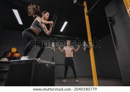Two young crossfit athletes exercising together at gym. Sporty fit couple having interval functional training class. Pretty female doing box jump exercise while muscular male doing back squats with ba Royalty-Free Stock Photo #2040180779